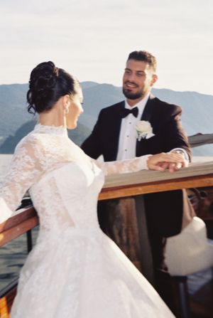 Bride in lace gown holds groom's hand on a classic wooden boat with Lake Como mountains in background.