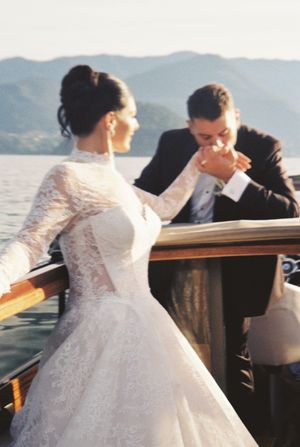 Groom in black tuxedo kissing bride's hand on a wooden boat with scenic mountain lake in background.