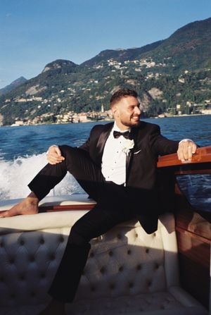 Groom in black tuxedo relaxing on a wooden boat on an Italian lake.