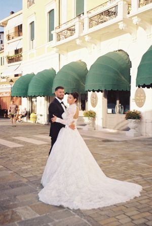 Bride and groom pose in elegant wedding attire outside a historic Italian building with green awnings.