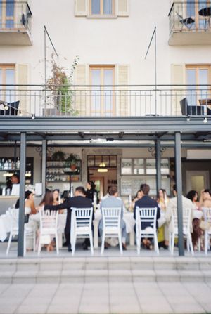 Wedding guests seated at white Chiavari chairs on outdoor restaurant terrace during daytime reception.