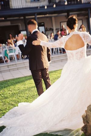 Bride in lace gown with open back walks with groom toward wedding guests at outdoor reception.