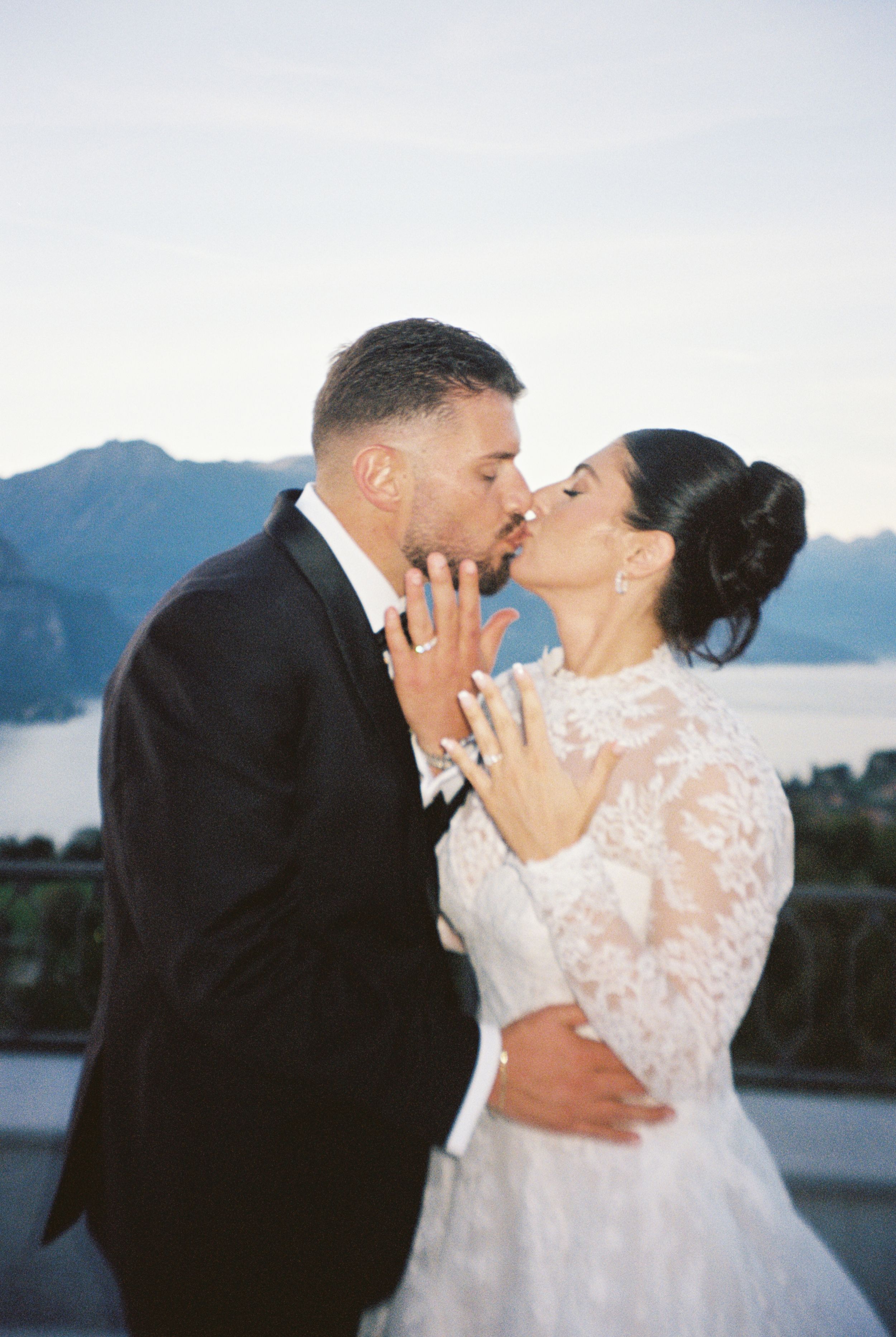 Bride and groom kissing on a terrace with mountain lake views, showing wedding rings, bride in lace gown, groom in black tuxedo.