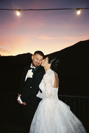 Bride and groom share a romantic kiss at sunset with string lights and mountain silhouettes in the background.