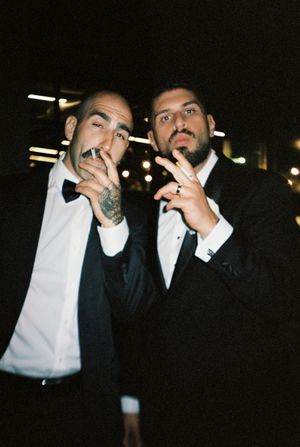 Two men in tuxedos posing at night, one with neck tattoos smoking a cigarette, both making hand gestures.