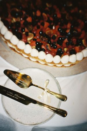 Elegant wedding cake topped with fresh berries and cream, displayed with gold engraved cake server and knife set.