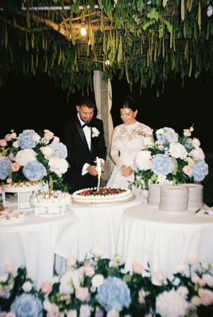 Bride and groom cutting wedding cake surrounded by blue hydrangea floral arrangements under hanging greenery.