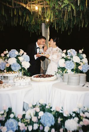 Bride and groom share cake at elegant wedding reception with blue hydrangea floral arrangements and hanging greenery backdrop.