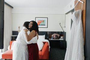 Bride and bridesmaid share an emotional hug in a hotel room with a white wedding dress hanging nearby.