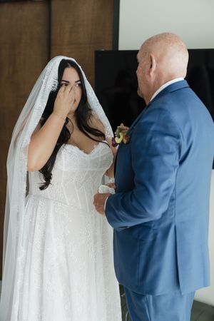 Emotional bride in white lace gown and veil covering her mouth while sharing a tender moment with man in blue suit.