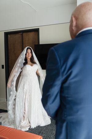 Bride in lace wedding gown and veil smiles at groom in blue suit during romantic first look moment.