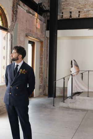 Groom in navy suit with floral boutonniere stands in industrial venue as bride descends staircase in background.