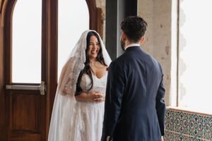 Bride in white gown and veil smiling at groom in navy suit during wedding ceremony near ornate tiled wall.