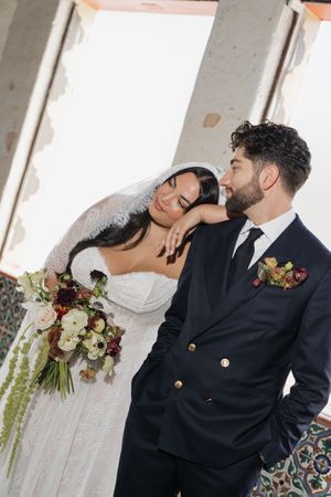 Bride leans on groom's shoulder holding burgundy bouquet, both in elegant wedding attire indoors.