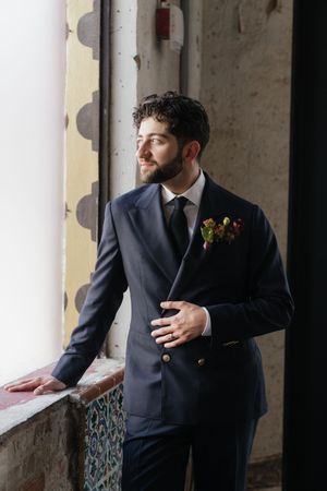 Groom in navy suit with floral boutonniere stands by window, looking sideways in elegant wedding attire.