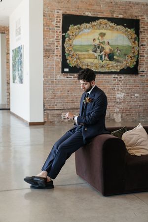 Groom in navy double-breasted suit sits on velvet sofa reading a note in a brick-walled wedding venue.