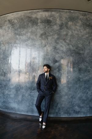 A groom in a navy suit with boutonniere leans against a textured gray curved wall, hands in pockets, looking away.