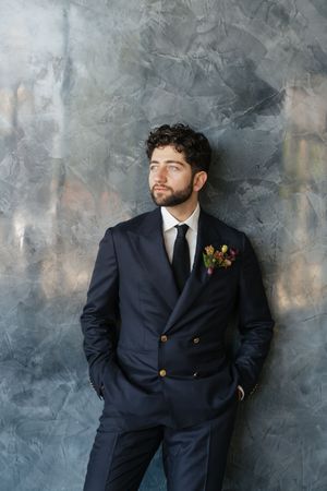 A dapper groom in a navy double-breasted suit with a colorful boutonniere stands against a gray textured wall.