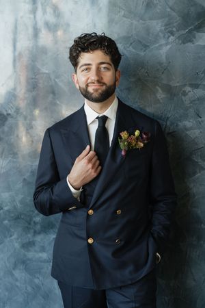 A stylish groom in a navy double-breasted suit with gold buttons, black tie, and floral boutonniere poses confidently.