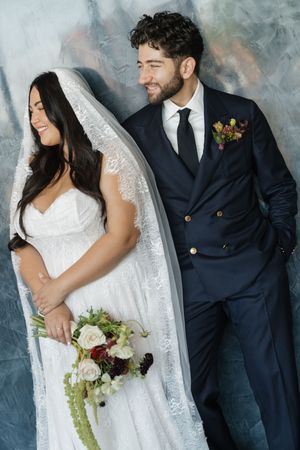 A smiling bride in a white lace gown and veil holds a bouquet beside a groom in a navy suit with a floral boutonniere.