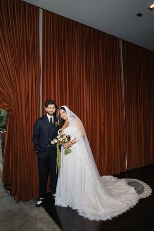 Bride in white lace gown with veil holds bouquet beside groom in navy suit before rust-colored drapes.