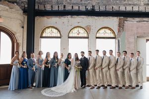 Wedding party poses indoors with bridesmaids in blue gowns and groomsmen in tan suits flanking bride and groom.