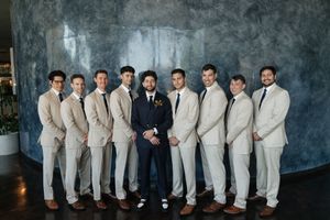 Groom in navy suit stands centered with nine groomsmen in matching tan suits, posing formally against a textured blue backdrop.