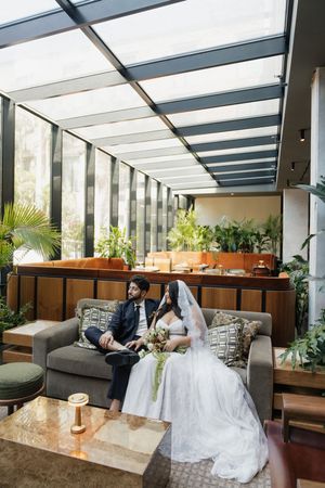 Bride and groom sit together on a gray sofa in an elegant glass-roofed venue surrounded by lush greenery.