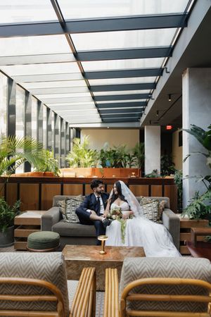 Bride and groom sit together on a gray sofa in an elegant indoor venue with lush greenery and a glass ceiling.