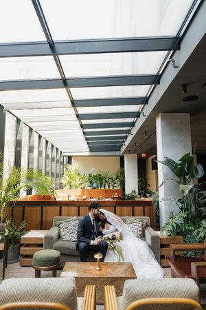 Bride and groom sit together on a sofa in an elegant indoor atrium venue filled with lush greenery on their wedding day.