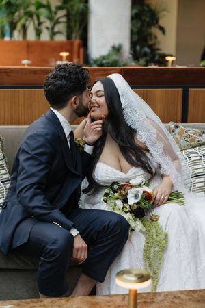 Bride and groom share a romantic moment on a couch, groom kissing bride's cheek as she smiles holding a bouquet.
