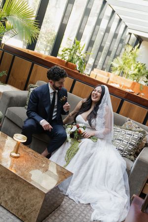 Bride and groom laughing together on a gray sofa in an elegant indoor venue with lush greenery.
