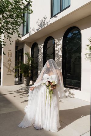 Bride in white lace gown and veil holds bouquet outside Haya hotel on sunny day.