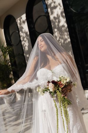 Bride in white lace gown and flowing veil holds cascading floral bouquet outdoors in sunlight.