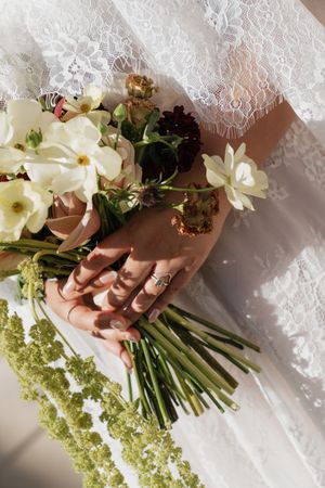 Bride holding a wedding bouquet of white and burgundy flowers with green stems, wearing a lace dress and diamond ring.