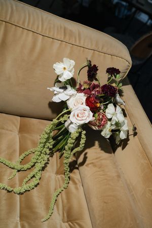 A romantic bridal bouquet with white, blush, and burgundy flowers and trailing green amaranthus on a gold velvet sofa.