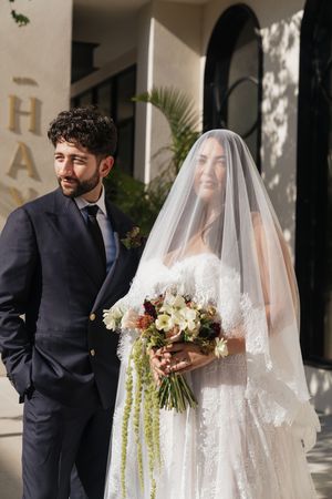Bride in lace gown and veil stands with groom in navy suit holding bouquet outside.