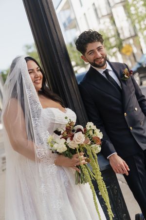 Smiling bride in white lace gown holding bouquet stands beside groom in navy suit near a black pillar outdoors.