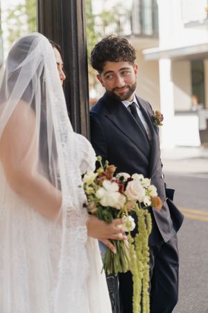 Groom in navy suit smiling at bride in white lace dress and veil holding a bouquet of flowers.