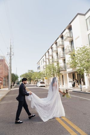 Bride and groom cross a sunny urban street, her lace gown trailing as he carries the veil train.