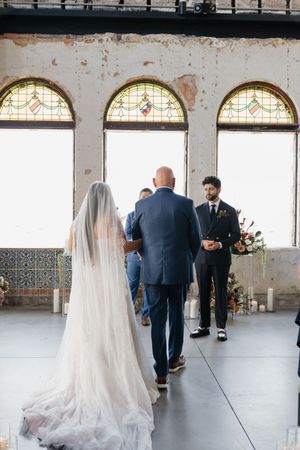 Bride walking down the aisle with escort toward groom in elegant venue with stained glass windows.