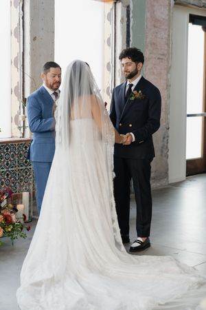 Bride and groom holding hands at indoor wedding ceremony with officiant, bride wearing long veil.