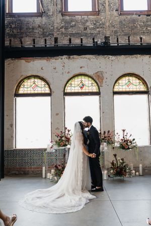 Bride and groom share first kiss at elegant wedding ceremony with stained glass windows and floral arrangements.