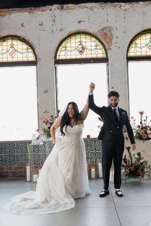 Bride and groom raise hands in celebration inside a rustic venue with stained glass arched windows and floral arrangements.