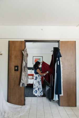 Woman in floral skirt selecting red dress from garment rack in hotel room doorway during wedding preparations.