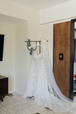 White wedding dress with veil hanging on wall hook beside teal heels in a bright hotel room near wooden door.