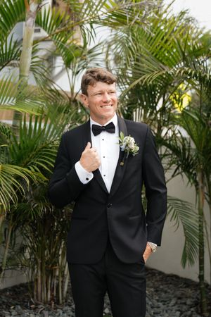 A groom in a classic black tuxedo and bowtie poses with a boutonniere against a tropical palm backdrop.