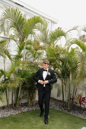 A person in a black tuxedo poses elegantly against lush tropical palm trees in an outdoor garden setting.
