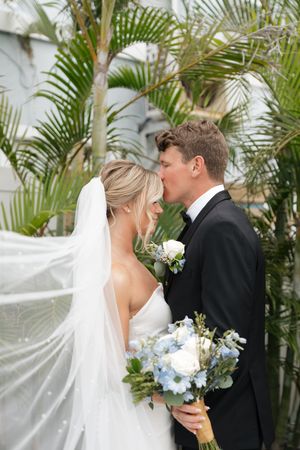 A newlywed couple shares a romantic moment among tropical palm fronds while the bride holds a blue and white bouquet.