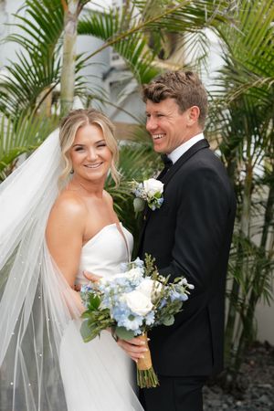 A bride in white gown holds blue bouquet next to groom in black tuxedo with tropical palm backdrop.